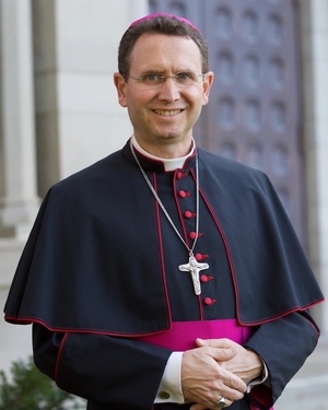 Bishop Andrew Cozzens, a smiling man in eyeglasses, dressed in a black tunic and short cape with fuschia piping and buttons and a cross hanging from a chain around his neck.